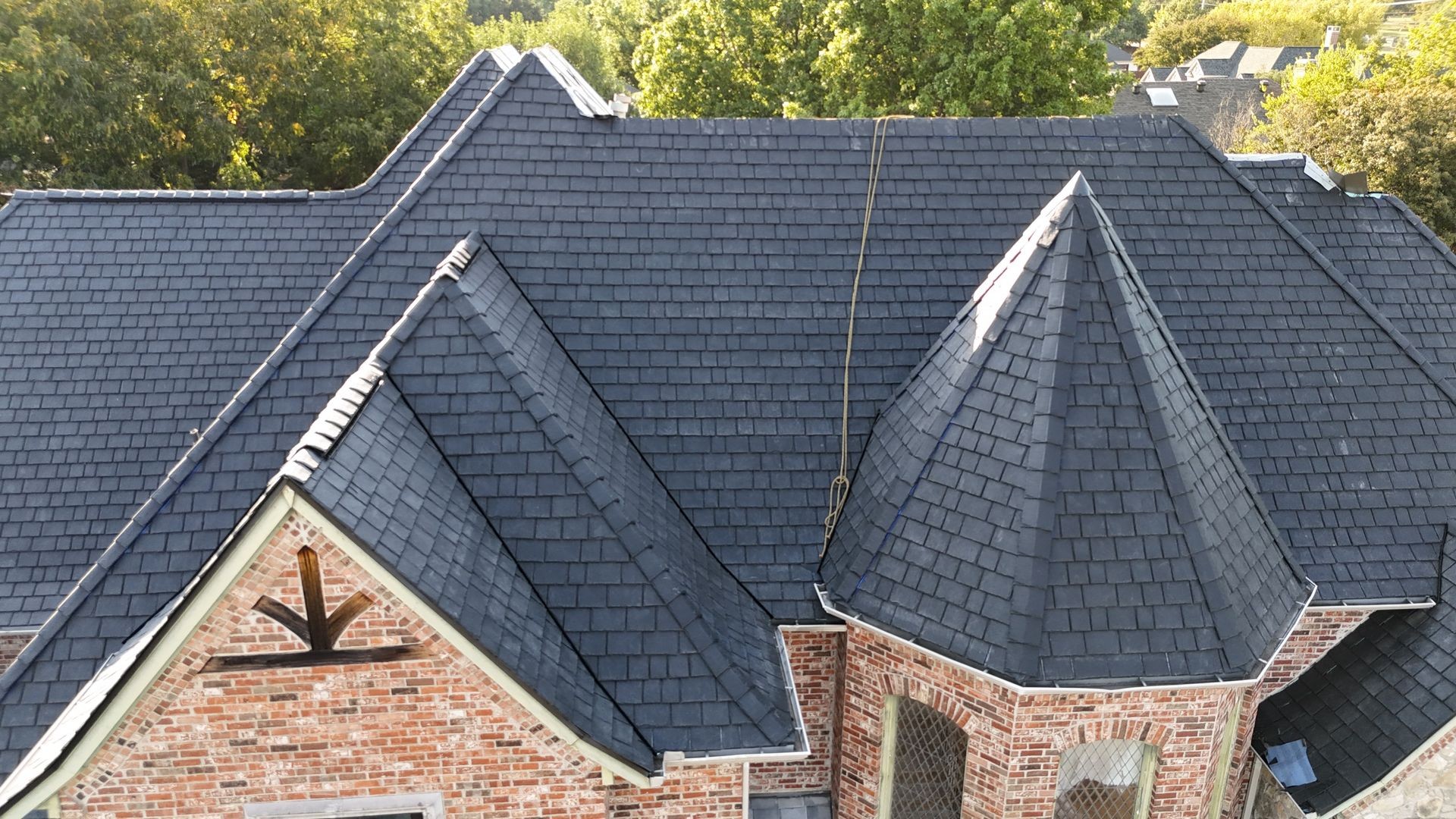 Aerial view of a house with dark slate roof tiles and multiple peaks surrounded by green trees.