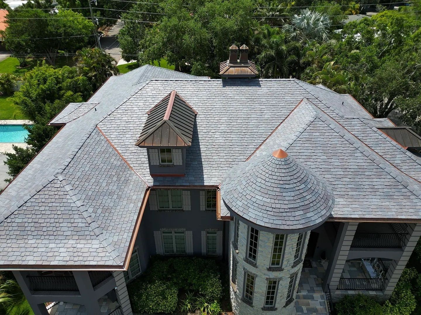 Aerial view of a large house with a gray shingle roof and lush green surroundings.