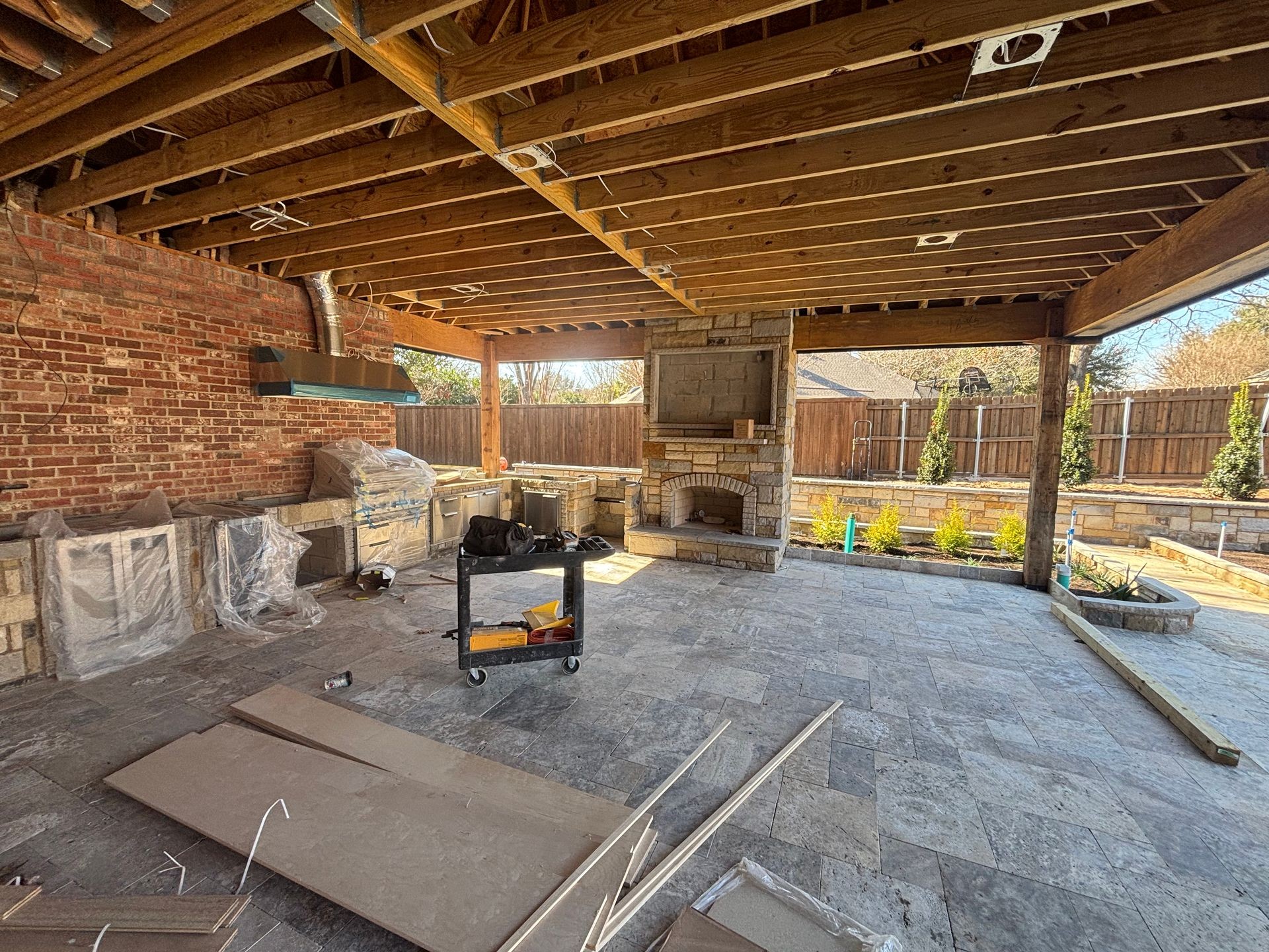 Outdoor patio under construction with exposed wooden beams, brick walls, and partially installed kitchen appliances.