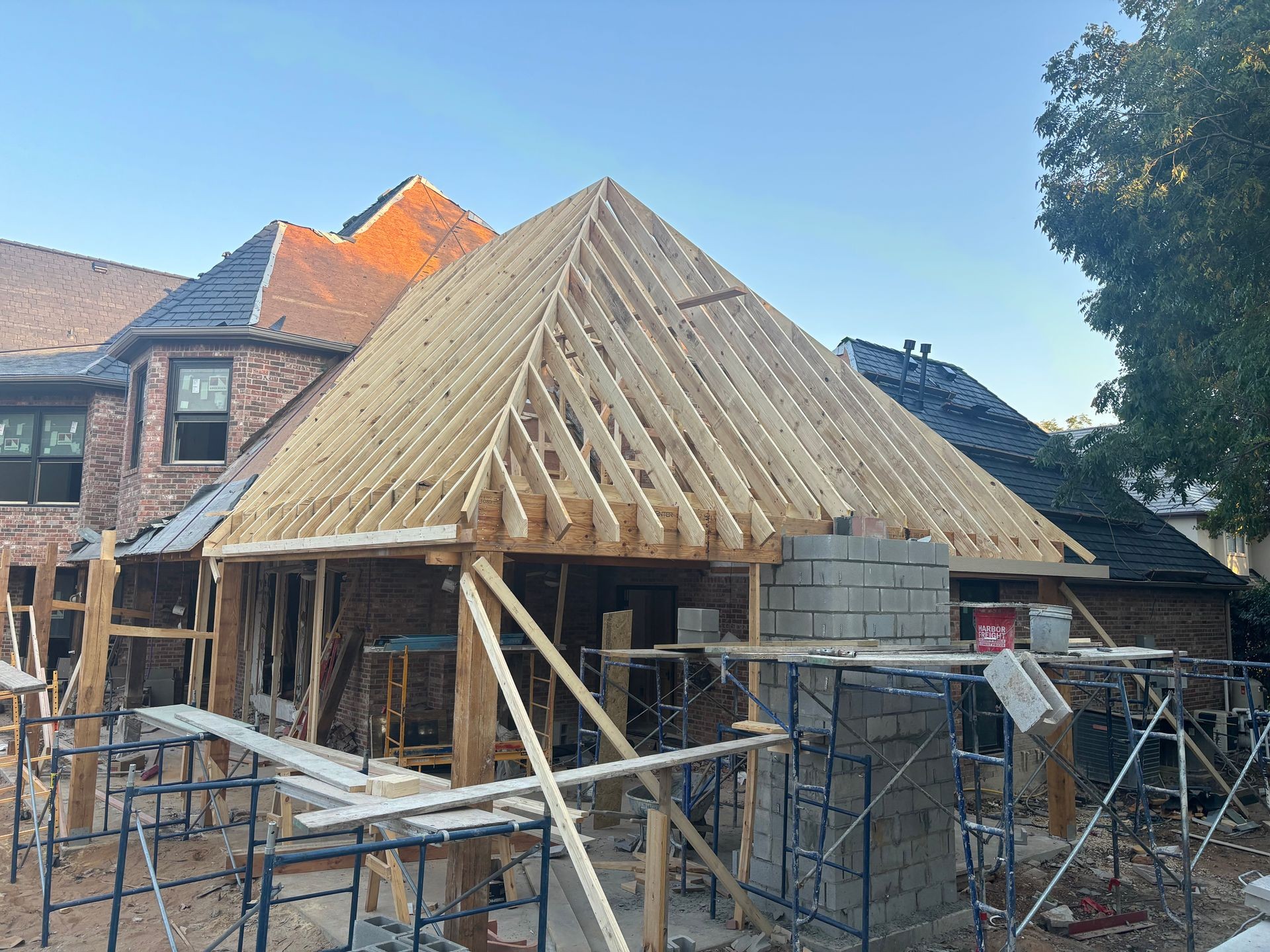 House under construction with wooden roof framing and brick walls, surrounded by scaffolding.