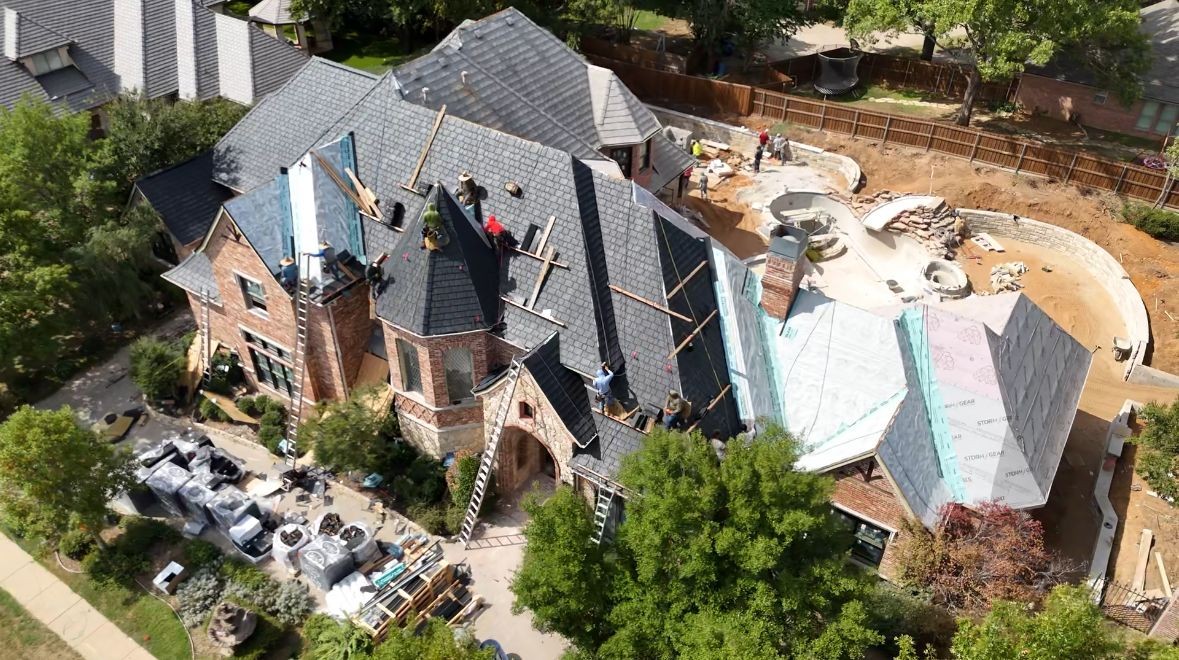 Aerial view of workers installing shingles on a large house under construction with landscaping materials nearby.