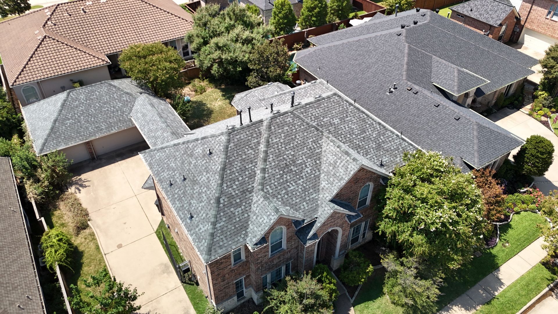 Aerial view of suburban houses with gray and brown roofs, surrounded by trees and driveways.